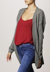 Woman wearing a grey knitted cardigan, red sleeveless top, and blue jeans, posing with hands relaxed by sides.