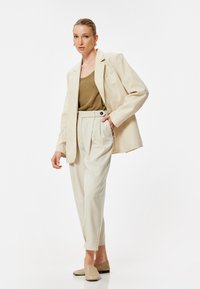 Blazer beige tagliato sopra una canotta marrone, abbinato a pantaloni crema a vita alta. Tessuto morbido e vestibilità ampia, completato con scarpe slip-on beige.