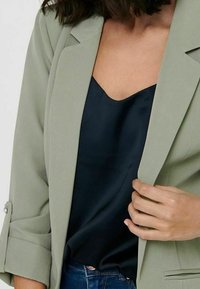 Eine Frau trägt ein hellgrünes Blazer über einem schwarzen Satin-Camisole mit hochgekrempelten Ärmeln und blauen Jeans, während sie den Revers des Blazers hält.