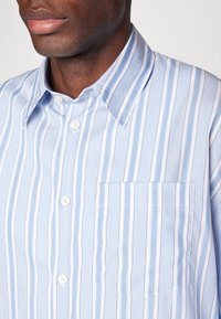 Camisa de botones a rayas azul claro y blanca con un único bolsillo en el pecho, cuello clásico y tejido texturizado. Presenta pequeños botones blancos.
