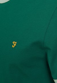 Green cotton t-shirt featuring a ribbed texture and rounded neckline, with an embroidered yellow logo on the left chest area.