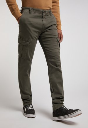 Mężczyzna w oliwkowozielonych spodniach typu cargo, czarnych trampkach z białymi sznurowadłami oraz brązowej bluzce z długim rękawem, stojący na jednolitym tle.