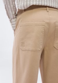 Μπεζ παντελόνι με μεγάλες πίσω τσέπες, μία από τις οποίες δείχνει μια μικρή ετικέτα "Margain Sports", φορεμένο με ένα ανοιχτό ριγέ πουκάμισο που είναι κρυμμένο μέσα.