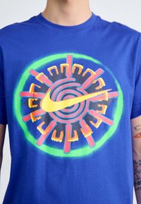 Modré bavlněné tričko s velkým vícebarevným kruhovým motivem se žlutým logem Nike ve tvaru zaškrtnutí, obklopeným červenými a zelenými akcenty.