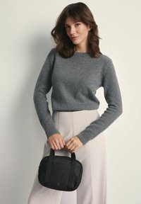 Czarna torba materiałowa z krótkimi uchwytami; prostokątny kształt. Modelka ma na sobie szary sweter z dzianiny i beżowe spodnie o wysokim stanie. Gładka tekstura.