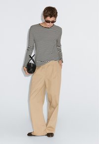 Gestreiftes Langarmhemd in Schwarz und Weiß, beige weit geschnittene Hose, hält eine kleine geflochtene schwarze Handtasche, trägt dunkle Sonnenbrille.