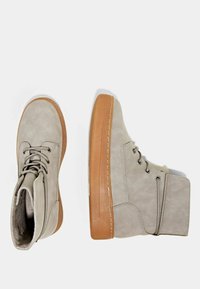 Bottines en daim gris avec une semelle en caoutchouc marron clair, dotées de six œillets ronds et de coutures décoratives le long du côté et de la base.