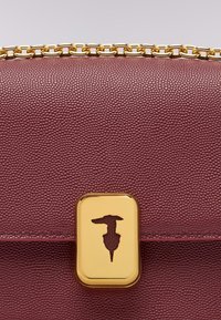 Burgundská kožená taška s texturovaným povrchem, zlatými doplňky, řetízkem na rameno a zlatým zapínáním s vyřezávaným designem.