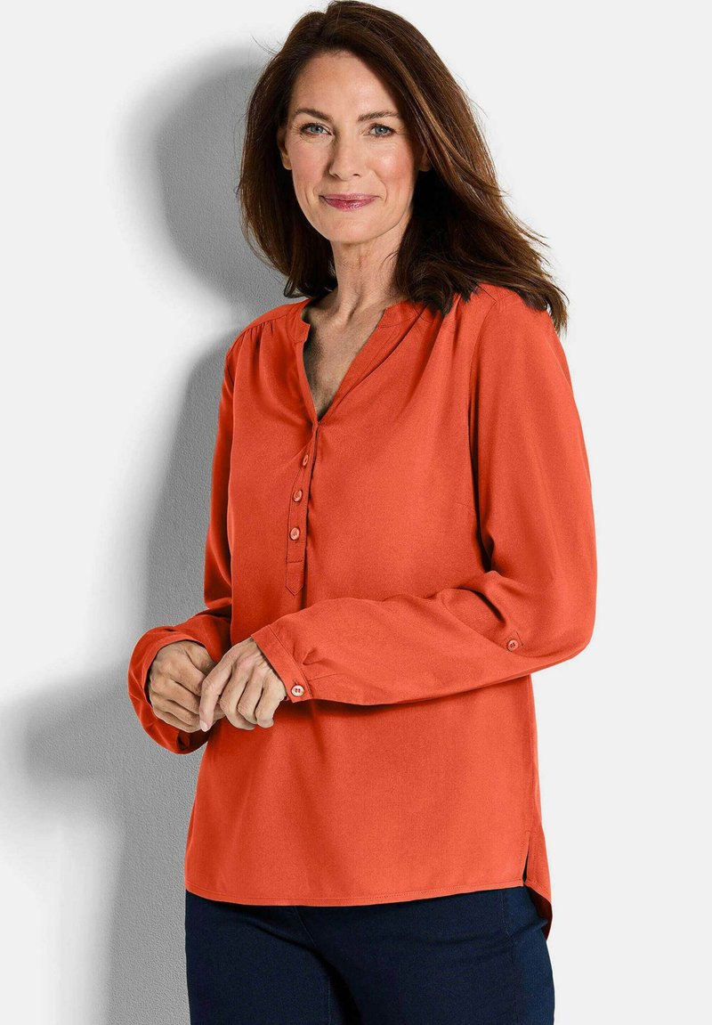 Orange Bluse mit langen Ärmeln, V-Ausschnitt und Knopfleiste. Weicher Stoff mit glatter Textur, mit seitlichen Schlitzen am Saum.