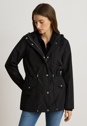 Donna che indossa una giacca nera con cappuccio, tasche frontali e coulisse, abbinata a jeans blu, guarda verso il basso con una mano dietro la schiena.