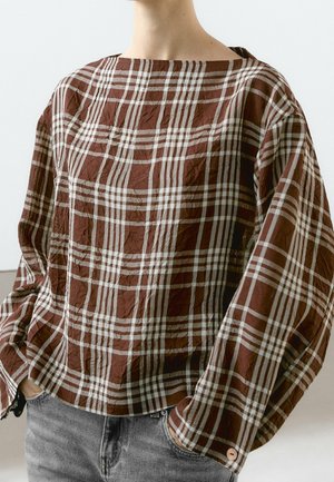 Person wearing a brown and white plaid long-sleeve shirt with a loose fit and hands in gray jeans pockets.