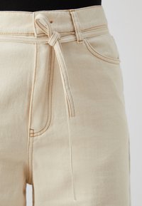Beige denimbukser med en knyttet sløyfe i midjen, synlige sømdetaljer og frontlommer. Glatt tekstur og skreddersydd passform.