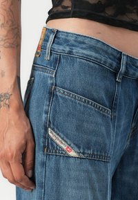 Jean en denim de couleur bleue moyenne avec une texture délavée, comportant une étiquette sur la poche arrière et plusieurs poches, cousues avec un fil contrasté.