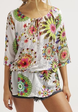 Woman wearing a white blouse and shorts with colorful floral and circular patterns, featuring a tie at the neckline and three-quarter sleeves.