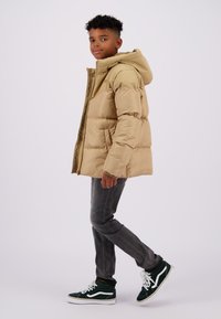 Pufferjack in lichtbeige, met een capuchon, ritssluiting en gestikte secties. Gecombineerd met donkergrijze jeans en groene sneakers.