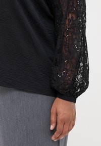 Černý top s texturovaným tělem a průhlednou černou krajkou, se třpytivými balónovými rukávy; přiléhavý manžet na zápěstí; vrstvený přes šedý kus oděvu.