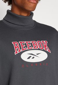 Šedá mikina s vysokým límcem, na které je umístěno velké červené logo "REEBOK" a bílé emblém. Měkká tkanina s hladkým povrchem a žebrovaným detailem límce.
