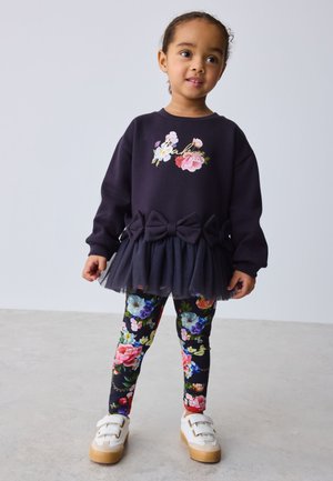 Niño pequeño de pie vistiendo una sudadera negra con estampado floral con lazos y tul, leggings florales y zapatillas blancas con velcro sobre un fondo liso.