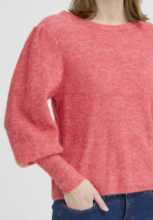 Kvinde iført en blød, lyserød langærmet trøje med puffede ærmer og blå jeans, hånd hvilende i lommen mod en ensfarvet baggrund.