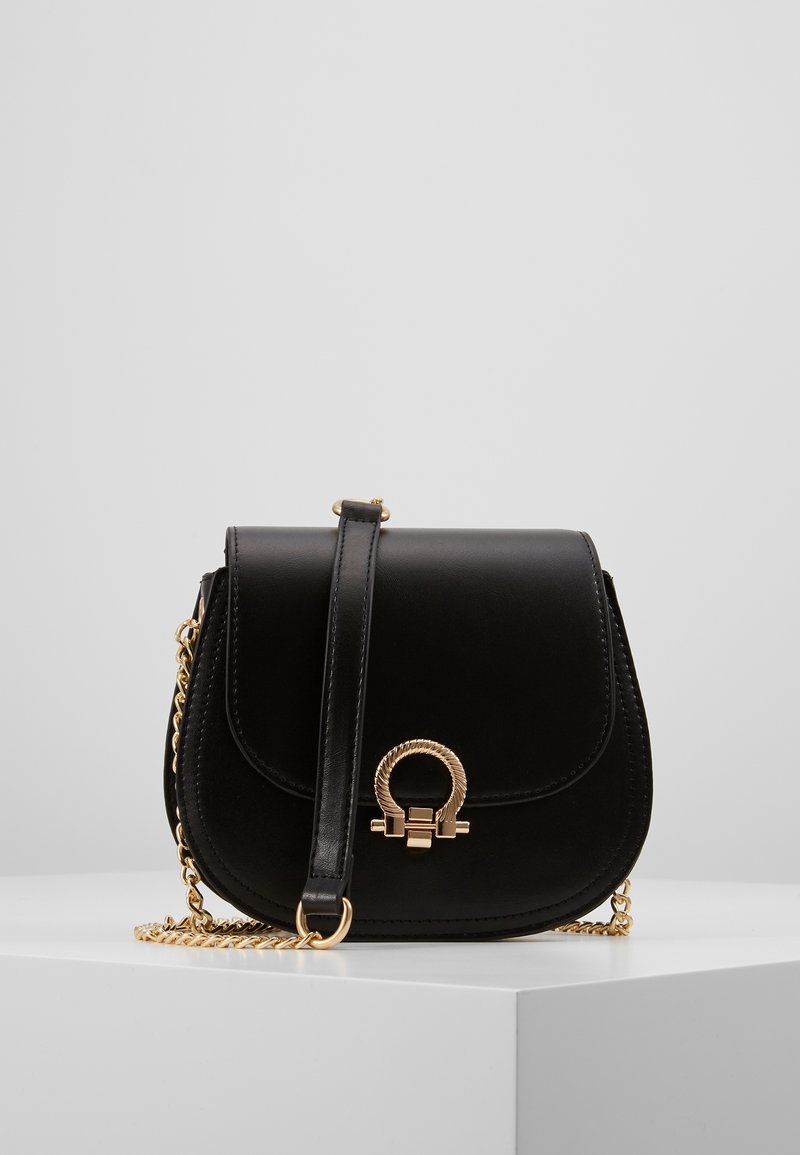 Borsa a tracolla in pelle nera con chiusura circolare dorata e tracolla a catena dorata, posata su un pedestal bianco contro uno sfondo grigio.
