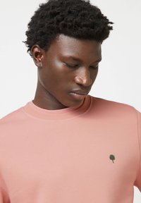 Felpa rosa chiaro con collo rotondo, dotata di un piccolo logo di un albero verde lato cuore. Textura liscia, linee pulite e design minimalista.