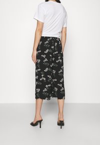 Pantalon culotte noir à motifs floraux blancs et verts, taille élastique, longueur mi-mollet, porté avec un t-shirt blanc uni.