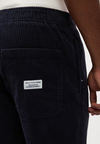Pantalons en velours côtelé foncé avec une poche arrière où se trouve une étiquette de marque visible. Tissu texturé avec un côtelage vertical. Taille élastique pour plus de confort.