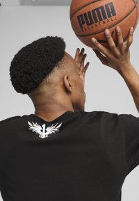Melna īsām piedurknēm T-krekls ar baltu grafiku, kas attēlo spārnus un numuru "1" aizmugurē. Valdītājs tur basketbola bumbu.