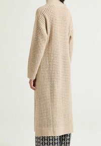 Maglione lungo beige lavorato a maglia con collo alto, motivo a rilievo e vestibilità ampia. Presenta spalle scese e dettagli a coste sull'orlo.