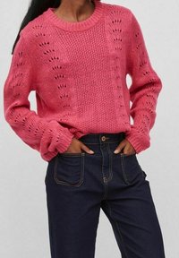 Maglione lavorato a maglia rosa con un motivo testurizzato, scollatura rotonda e maniche voluminose, abbinato a jeans alti di colore blu scuro.