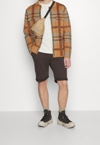 Plaid shirt in brown, orange, and gray; white t-shirt; brown shorts; beige fanny pack; gray and black sneakers with textured soles.