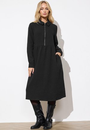 Vrouw draagt een zwarte midi-jurk met lange mouwen en een rits aan de voorkant, staand met één hand in de zak en zwarte laarzen tot halverwege de kuit.