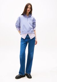 Sudadera de color púrpura claro con cuello alto, superpuesta a una camisa de cuadros, combinada con jeans de campana azules y zapatos negros. Fondo liso.