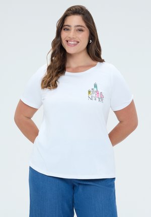 Mujer sonriente con cabello largo y ondulado lleva una camiseta blanca con un estampado colorido del skyline de Nueva York y vaqueros azules sobre un fondo liso.