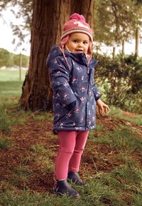 Palton căptușit de culoare bleumarin cu model floral, beanie roz cu pompon, colanți roz și cizme bleumarin. Stând pe iarbă lângă un copac.