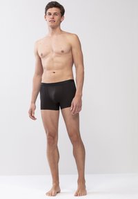 Boxer briefs noirs en tissu lisse avec une coupe ajustée, présentant un léger brillant et une ceinture douce, portés par un mannequin masculin se tenant debout.