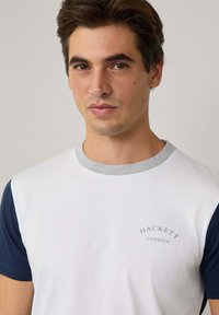 Camiseta de manga corta con cuerpo blanco, mangas azul marino y cuello gris. Presenta el logo "Hackett London" en letras azules.
