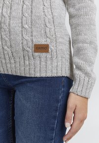 Gray knitted sweater featuring a cable design, ribbed cuffs, and a leather label with "OXMO" on the side. Paired with blue skinny jeans.