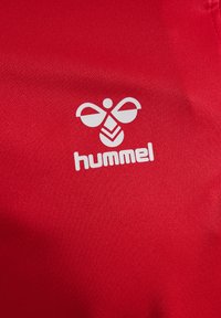 Roter Stoff mit glatter Textur zeigt ein auffälliges weißes Hummel-Logo, das ein stilisiertes Bienenmuster über dem Markennamen darstellt.