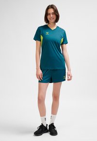 Ung kvinna stående, klädd i sportkläder i teal och lime med shorts, vita strumpor och svarta sneakers, leende mot en enfärgad bakgrund.