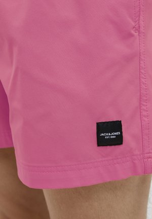 Bañador rosa con una textura suave, que presenta una etiqueta de tela negra con texto blanco en la parte inferior izquierda. Costuras visibles a lo largo del dobladillo.