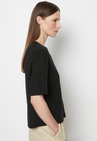 Schwarzes Strickoberteil mit kurzen Ärmeln, Rundhalsausschnitt, lässiger Passform und seitlichen Schlitzen am Saum. Der Stoff wirkt weich mit einer subtilen Textur.