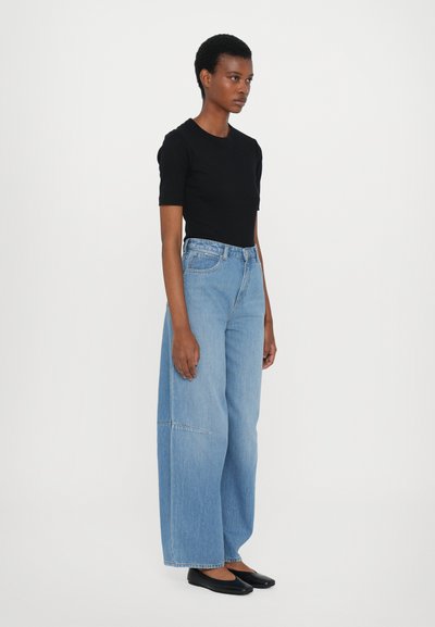 Schwarzes, kurzes, tailliertes Oberteil, breite, hellblau jeans mit einer Nahtdetails und schwarze flache Schuhe, stehend vor einem weißen Hintergrund.