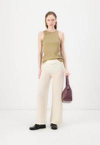 Olivově zelený top bez rukávů, bílé široké kalhoty s texturou a černé boty. Modelka drží malou fialovou kabelku, která ukazuje minimalistický design.