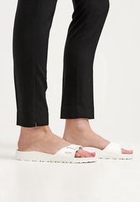 Pieds portant des sandales blanches à enfiler avec boucles, associées à un pantalon noir slim coupé au-dessus de la cheville.