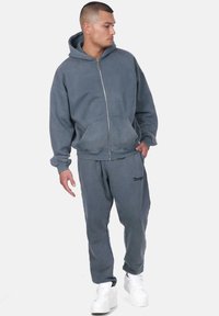 Hoodie zippé gris et jogging assorti, fabriqués en matériau doux. Comprend des poches avant, des poignets élastiques et un détail de logo discret. Baskets blanches.
