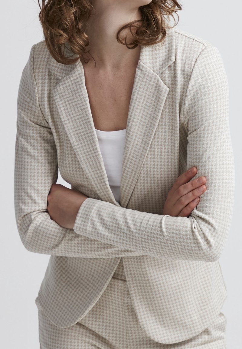 Femme aux cheveux bouclés portant un blazer beige à motif pied-de-poule et un pantalon assorti, bras croisés sur un haut blanc, sur un fond uni.