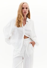 Weißes, strukturiertes Hemd mit Knöpfen, das über einer lockeren Hose mit Kordelzug getragen wird, beide aus einem leichten Stoff; hat einen lässigen Schnitt und lange Ärmel.