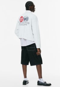 Grå cropped sweatshirt med blå og rød grafik, båret over en lys rosa skjorte med krave; parret med sorte denim shorts og sorte sko.