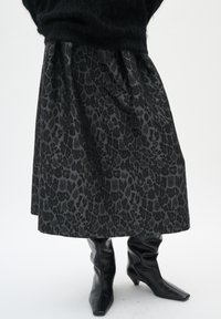 Schwarze Midirock mit dezentem dunklem Leopardenmuster, kombiniert mit schwarzen, quadratischen Leder-Mid-Heel-Stiefeln vor einem weißen Hintergrund.